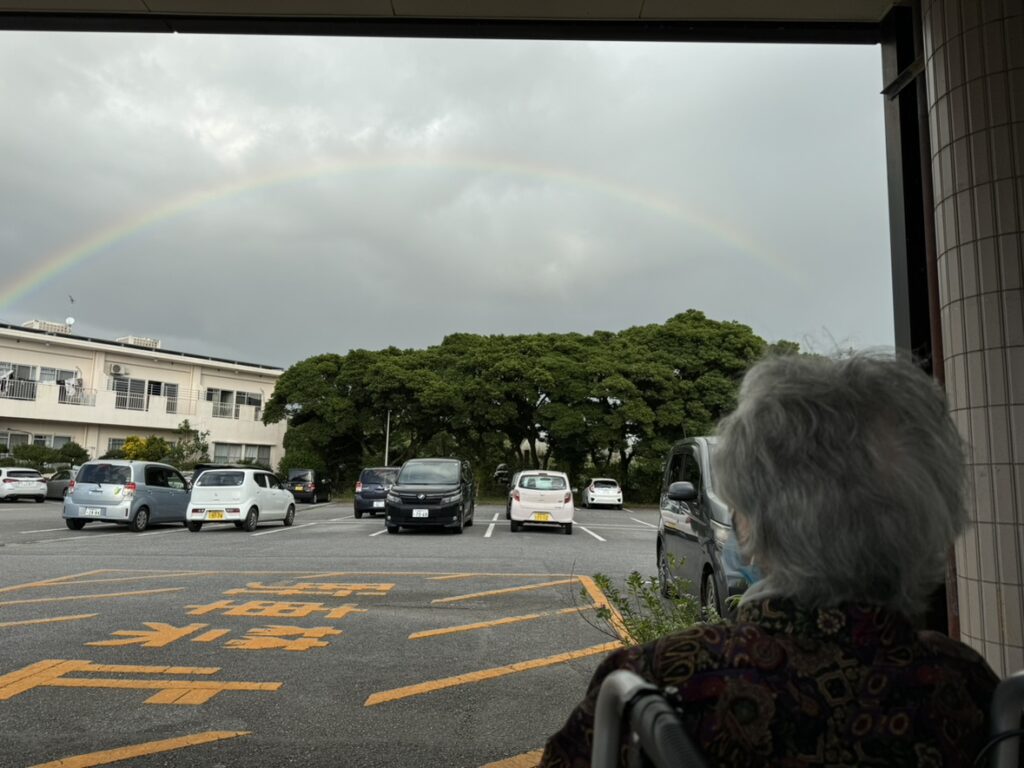 施設駐車場と周辺環境。虹が架かる空の下、駐車車両と大きな樹木が見える。
